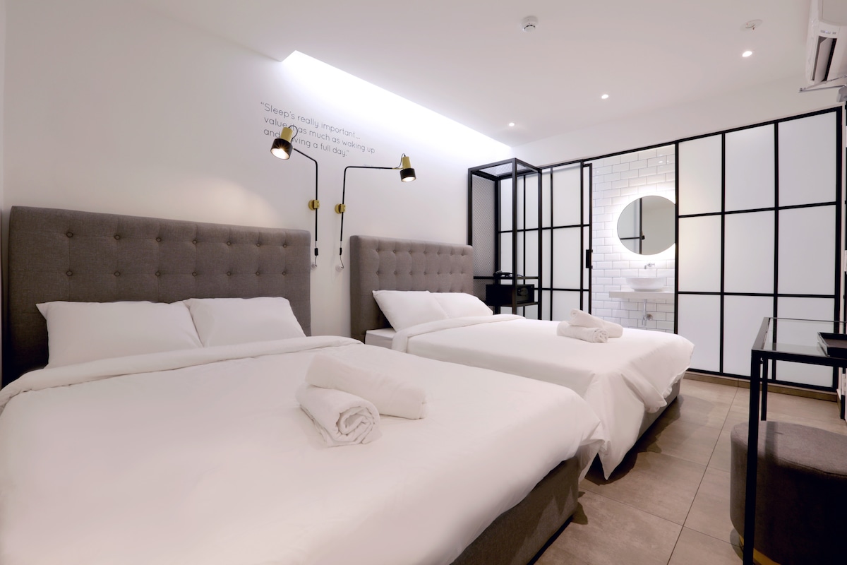 A clean and modern room features two spacious beds with neutral bedding, complemented by minimalistic decor. A round mirror and stylish lighting accentuate the white walls, while a black framed glass partition separates areas. A small desk is positioned against a wall, offering additional functionality.
