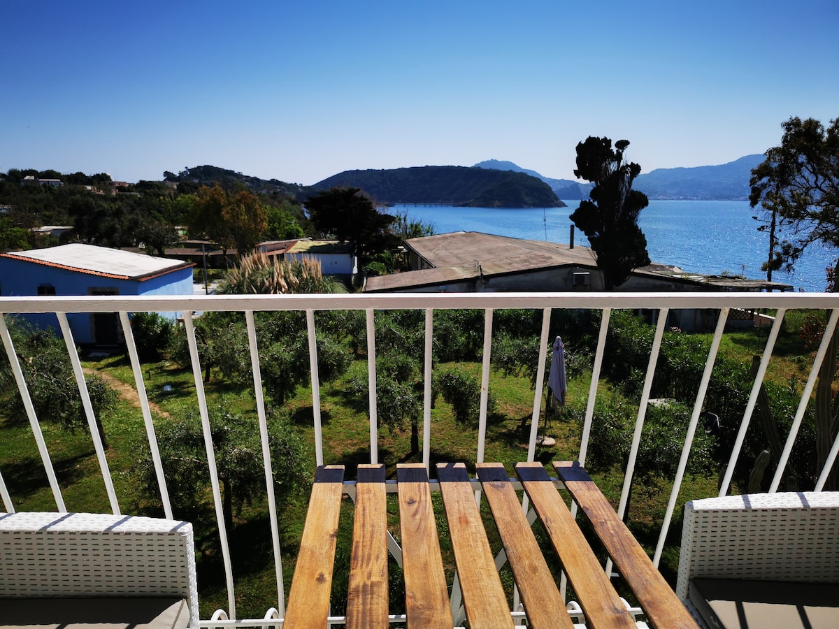 The balcony features wooden slats and overlooks a tranquil landscape with views of the sea and nearby hills. Lush greenery is visible below, enhancing the scenic backdrop and inviting relaxation in this outdoor setting.