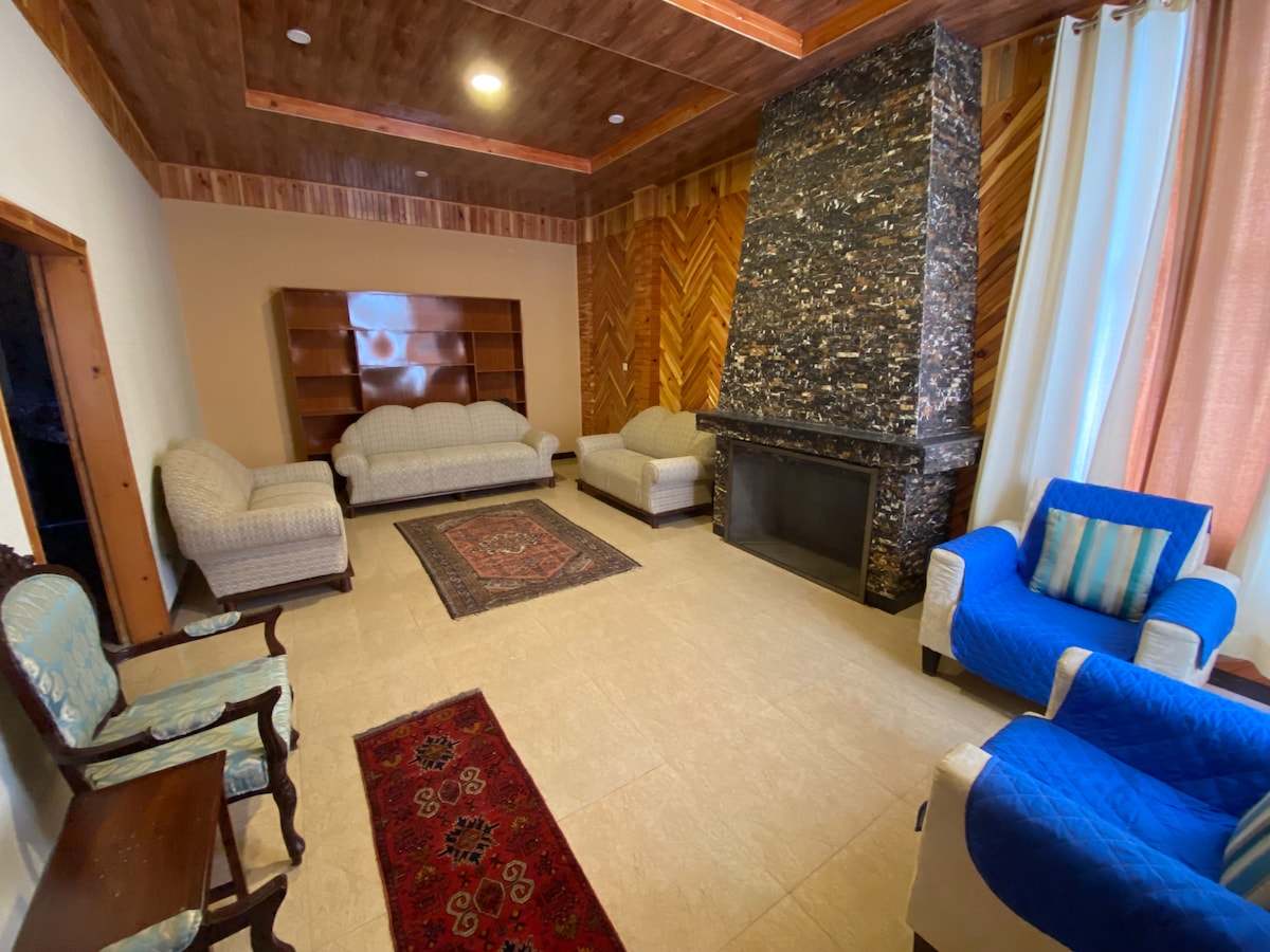 A spacious lounge area features a stone fireplace, flanked by two sets of seating arrangements. Two blue armchairs and two patterned sofas provide ample seating. Shelves along one wall offer space for decorative items. Natural light filters in through the large windows, complemented by light-colored drapes.