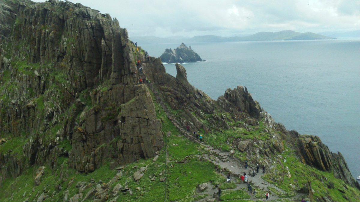 A rugged coastline is depicted with steep cliffs and rocky outcrops. The water below reflects shades of blue and gray, while distant islands are visible on the horizon. Several hikers can be seen navigating the terrain, highlighting the area's natural beauty and outdoor activity potential.