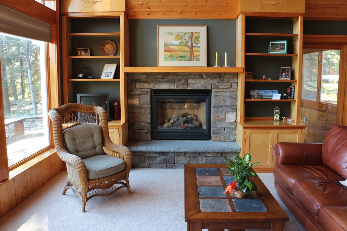 A welcoming living area features a stone-lined fireplace centered between built-in wooden shelving. A comfortable wicker chair and a leather sofa are positioned around a coffee table with a glass top. Natural light floods the space through large windows, enhancing the cozy ambiance.