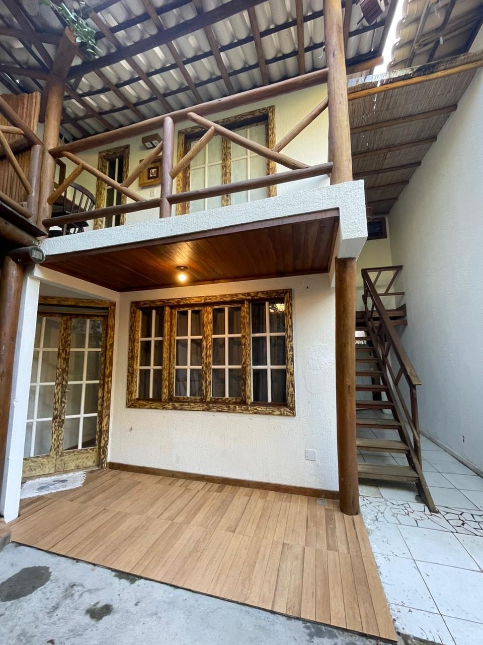 An outdoor area is presented with a wooden floor leading to a charming entrance, framed by large windows and intricate detailing. A staircase ascends to an upper level, while wooden beams and a sheltered roof provide a cozy ambiance.