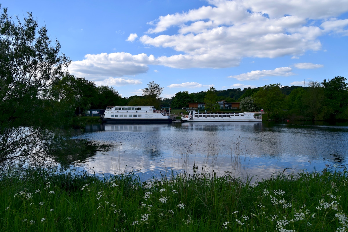 Two boats are moored on a serene river, surrounded by lush greenery. The calm water reflects the blue sky scattered with soft, fluffy clouds. Grass and wildflowers line the riverbank, enhancing the natural beauty of the landscape.