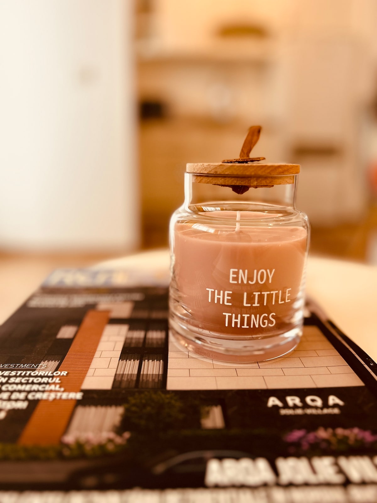 A softly lit jar candle sits atop a magazine, featuring a wooden lid and a light pink hue. The candle bears the inscription 'Enjoy the little things', creating a calming focal point in the space.
