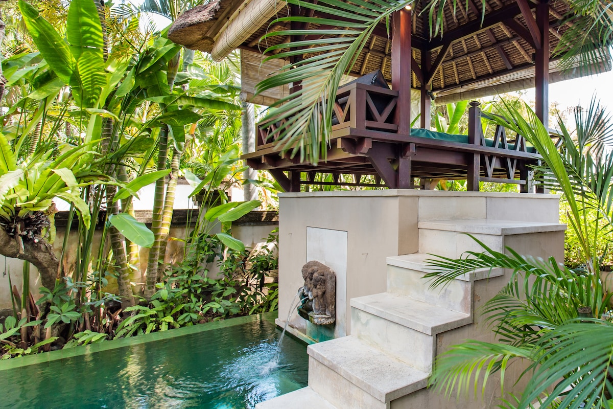 An elevated resting pavilion is surrounded by lush greenery, offering a space with a daybed. Steps lead down to a serene plunge pool, complemented by a stone water feature. The tropical setting enhances the tranquil ambiance of the area.