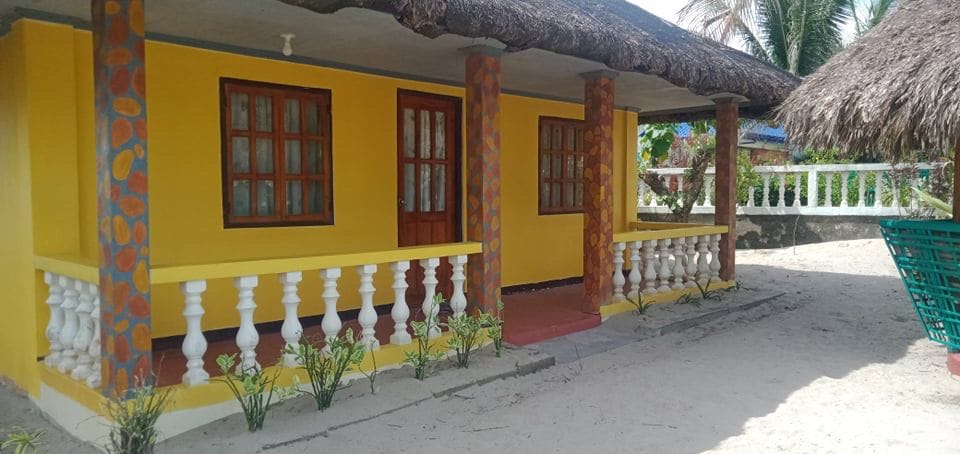 A vibrant yellow exterior of a home is showcased, framed by four columns with decorative stone patterns. Large windows with wooden frames provide natural light, while a sandy area surrounds the structure. Palm trees can be seen in the background, enhancing the tropical feel.