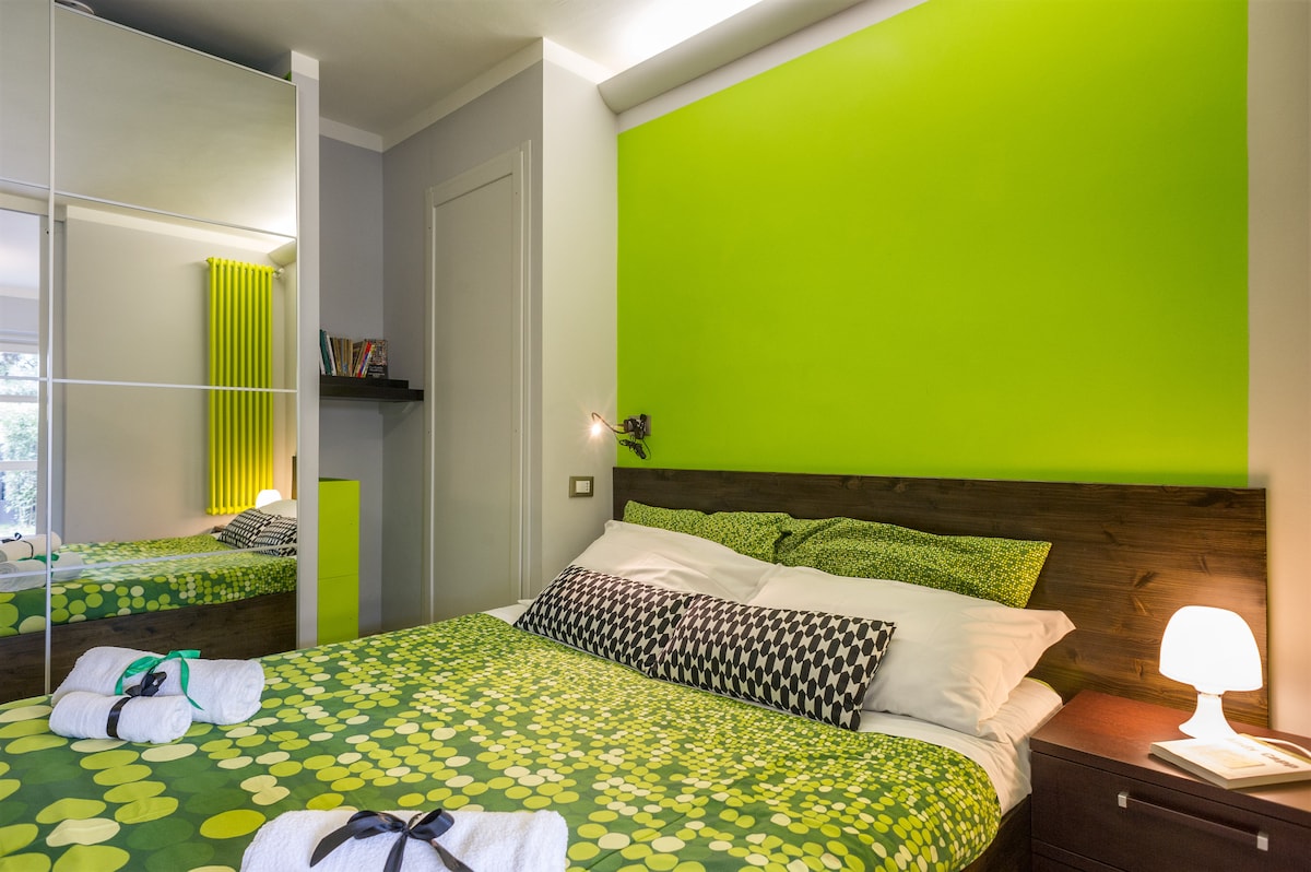 A vibrant sleeping area displays a green accent wall behind a spacious bed. The bed is adorned with patterned bedding and decorative pillows. Soft lighting is provided by a bedside lamp. A closet reflects the room's surroundings, contributing to a contemporary design.