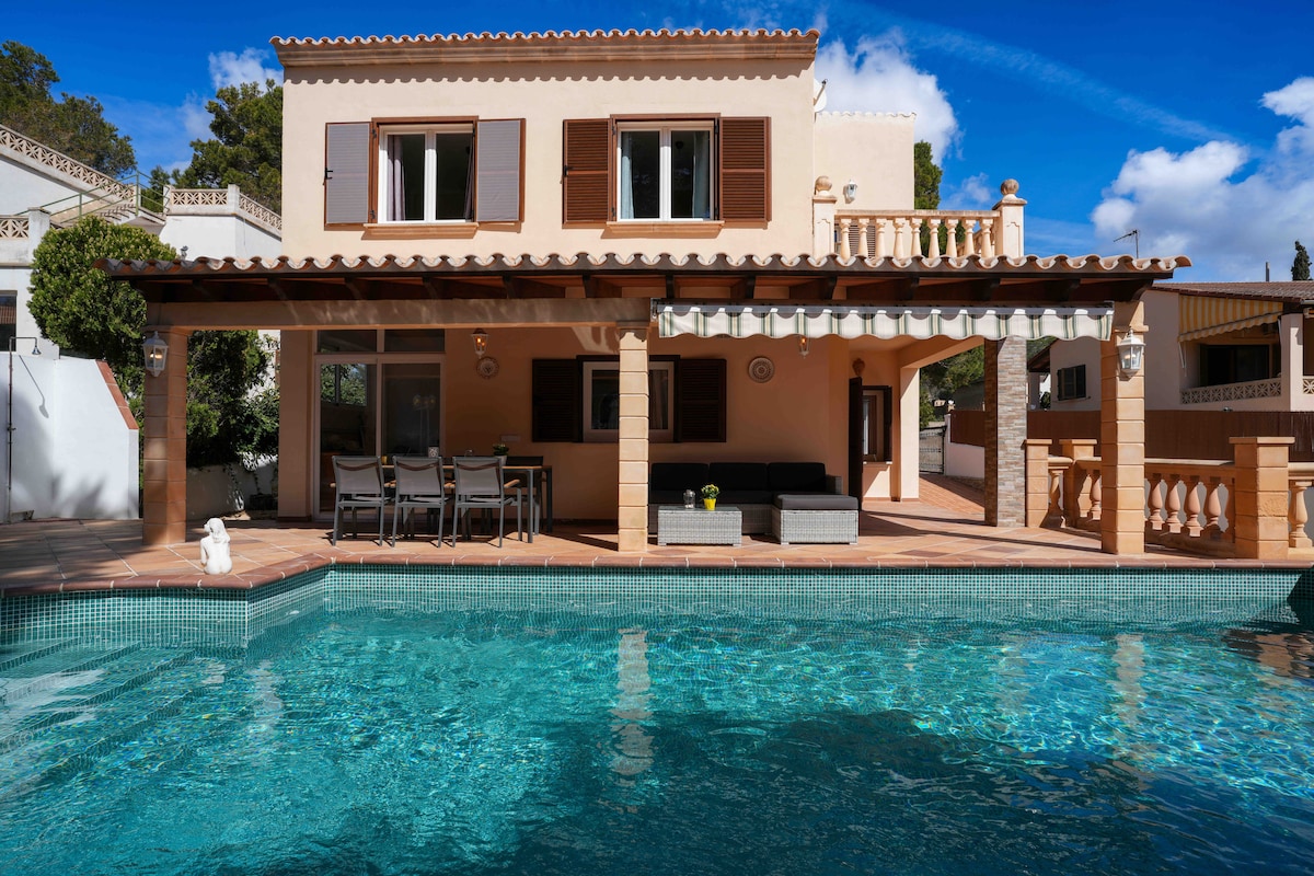 A view of the two-story house showcases a large outdoor terrace with dining furniture and a visible swimming pool. The pool reflects the sky, and the house is adorned with wooden shutters. Lush greenery surrounds the area, contributing to a serene environment.