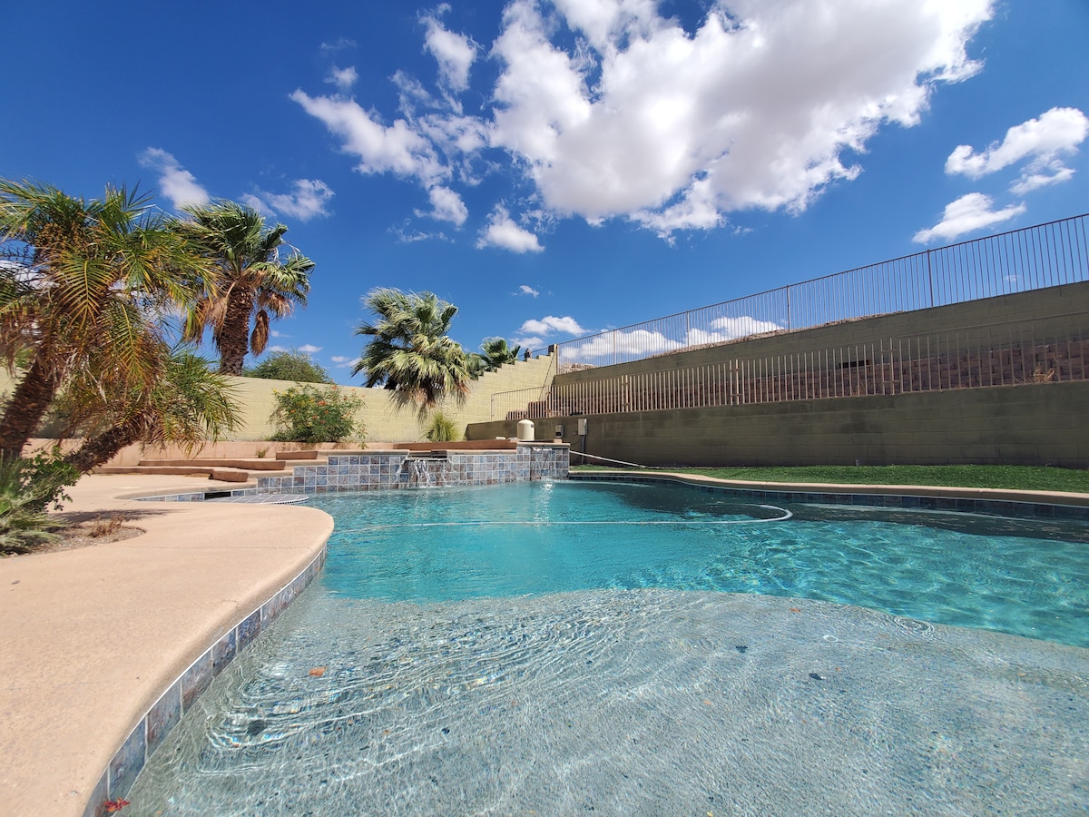 The backyard pool area features a refreshing pool under a clear blue sky dotted with fluffy clouds. Palm trees surround the space, and a low stone wall defines the perimeter, adding privacy to the serene environment.