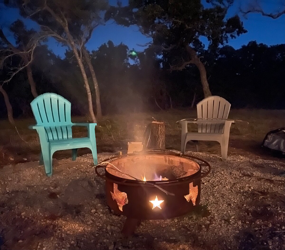A cozy fire pit takes center stage, surrounded by two Adirondack chairs—one in light blue and one in gray. Soft light from the flames illuminates the stone gravel beneath, with tree silhouettes gently framed in the evening sky.