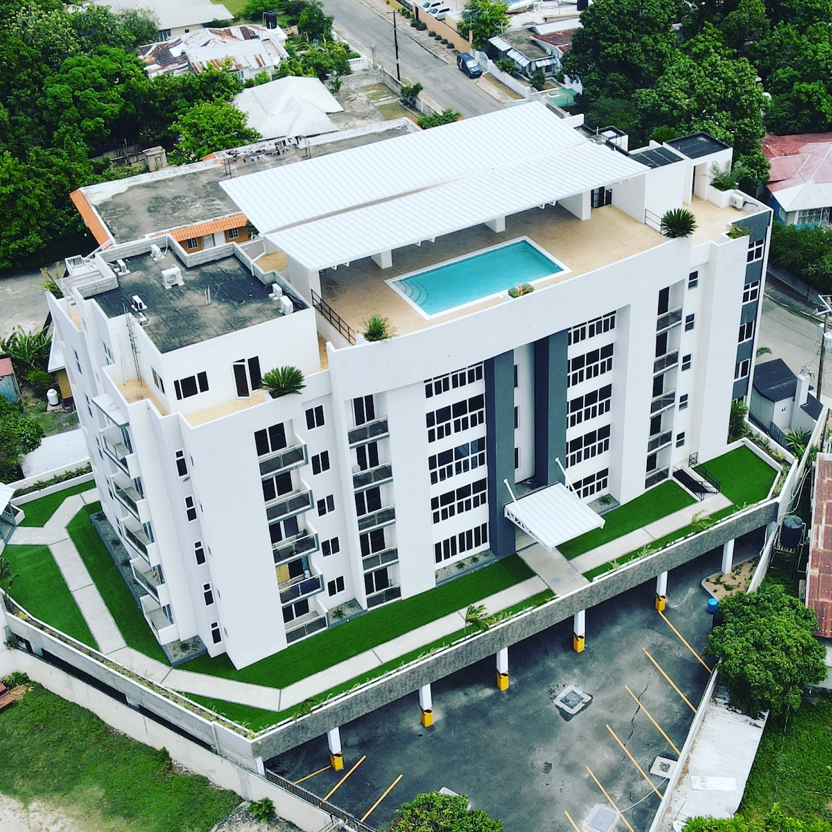 An aerial view captures a modern apartment complex with a rooftop pool surrounded by sun loungers. The building features a sleek design, with large windows and a grassy landscape below. Nearby, a parking area is visible, providing ample space for vehicles.