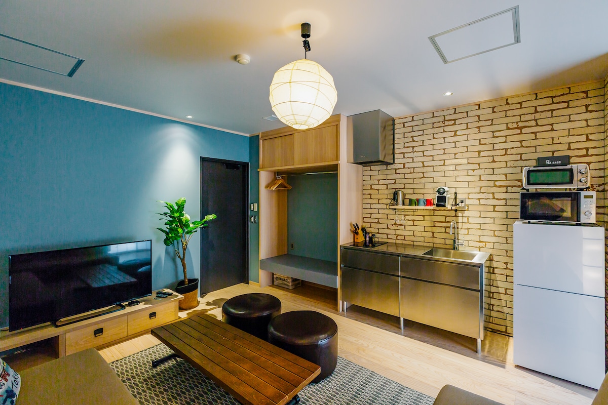 A modern living area is shown, featuring a sleek kitchen with stainless steel appliances and a decorative plant in the corner. A low wooden coffee table is placed in front of a comfortable seating area, which includes dark upholstered stools and a flat-screen television.