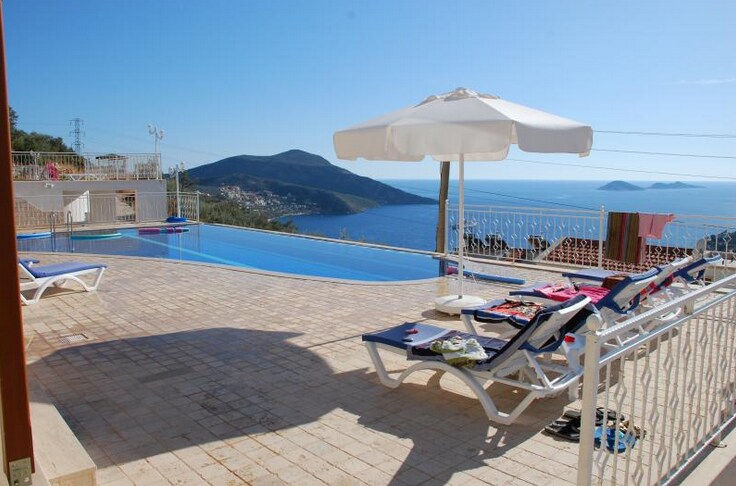 A spacious pool area features four sun loungers and a large umbrella providing shade. The infinity-style pool offers expansive views of the surrounding hills and coastline. Various colorful towels and swimwear are laid out on the loungers, highlighting the inviting atmosphere of the space.