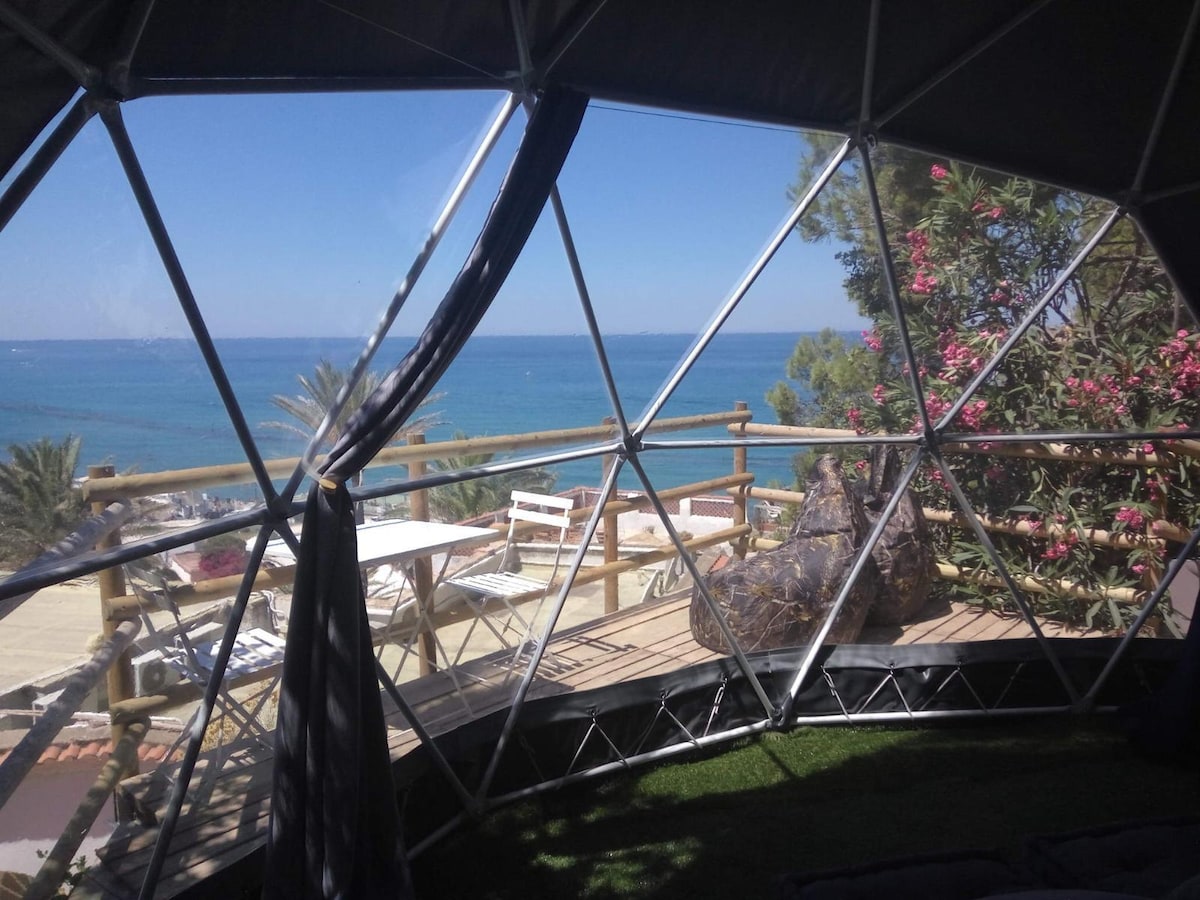 A geodesic dome features expansive clear panels that offer panoramic views of the ocean and surrounding greenery. A private terrace is visible, adorned with white furniture and surrounded by lush plants. The interior showcases a comfortable sleeping area, blending seamlessly with the breathtaking landscape.