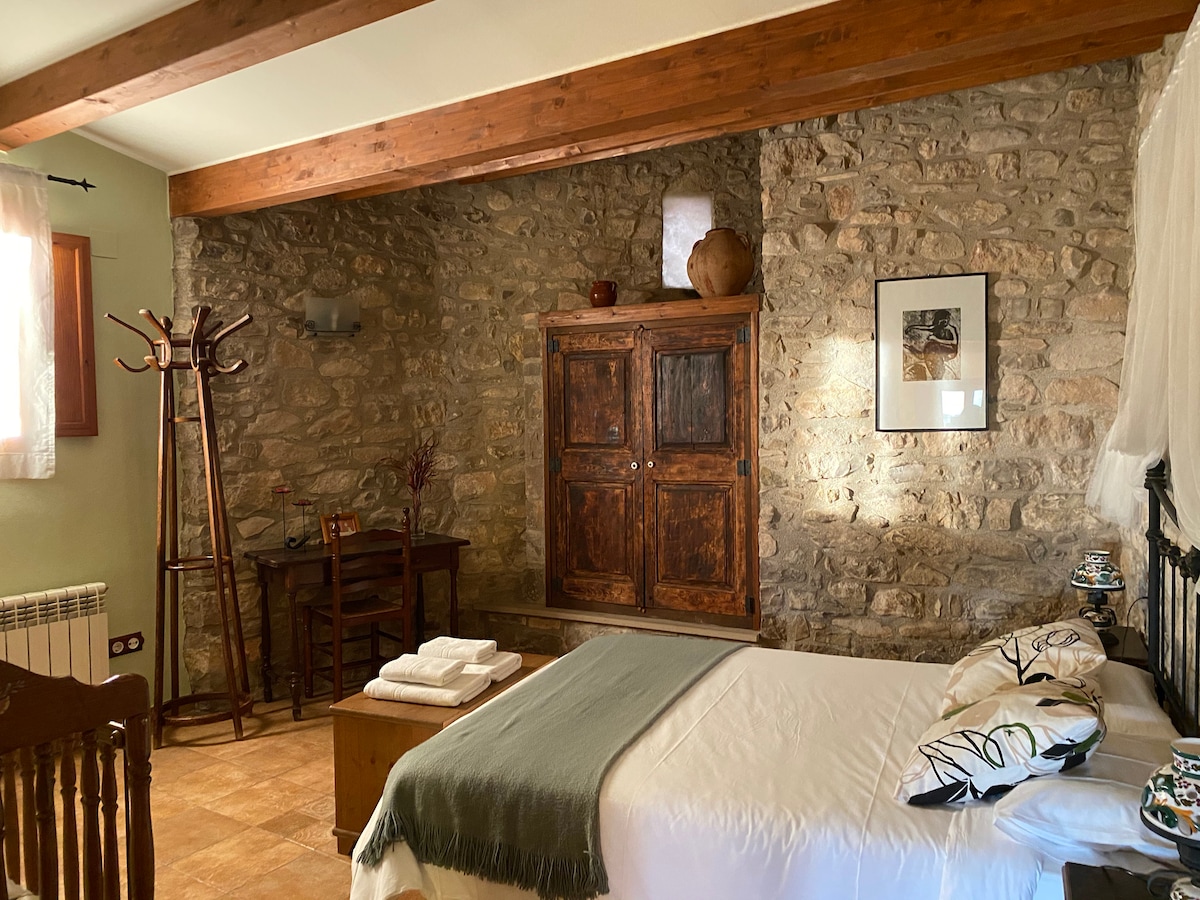 A rustic double bedroom features exposed stone walls complemented by wooden beams. A spacious bed is adorned with decorative pillows, and a soft throw lies at the foot. A small desk and coat rack are positioned nearby, creating a cozy, functional space.