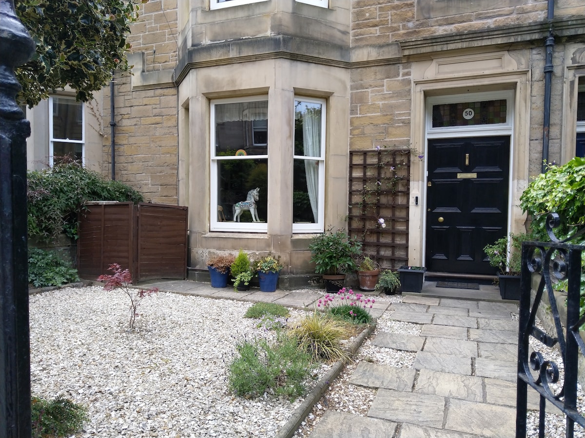 The exterior of a charming family home is depicted, featuring a stone façade and a black front door. A gravel garden is framed by potted plants and a small tree, adding greenery to the entrance. Large windows allow natural light to fill the interior.
