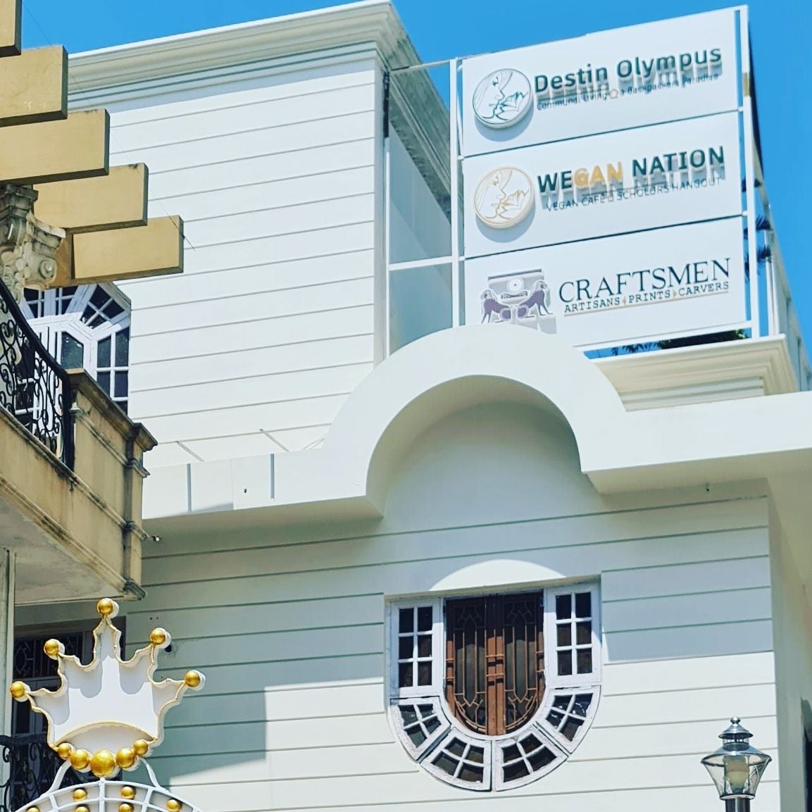The exterior of the building is shown, featuring a clean, modern facade with a combination of white and earthy tones. Signage is prominently displayed, including the names 'Destin Olympus,' 'Wegan Nation,' and 'Craftsmen.' Architectural details include arched windows and decorative accents.