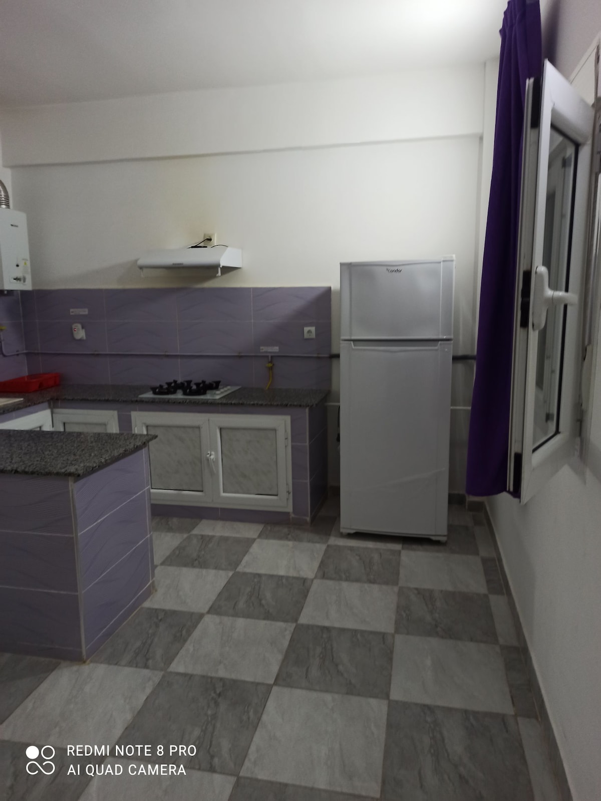 A functional kitchen space is equipped with modern appliances, including a white refrigerator and a gas stovetop. Light purple tile accents the walls, while granite countertops offer ample workspace. The flooring features a checkered pattern in neutral tones, and a window allows natural light in.