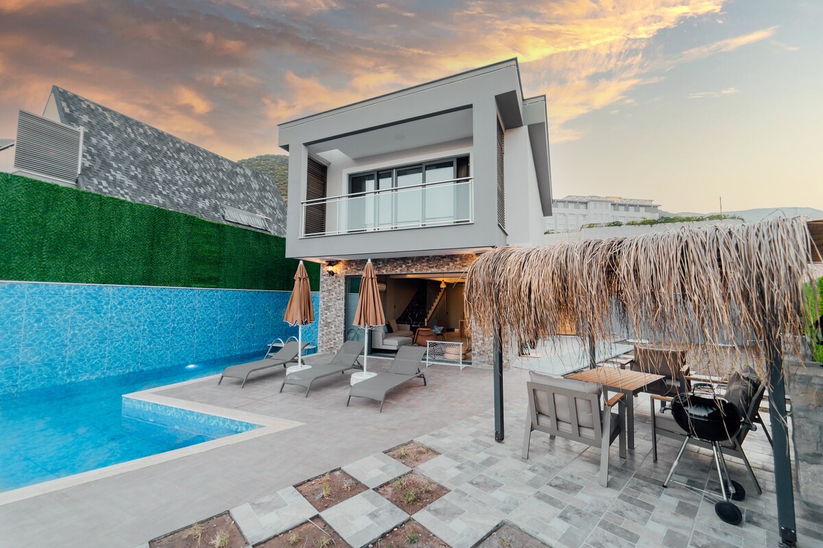 A modern villa exterior is presented, featuring a spacious swimming pool surrounded by lounge chairs and umbrellas. A covered dining area with a table and chairs is visible. The backdrop includes lush greenery and the building's clean architectural lines, under a colorful sunset sky.