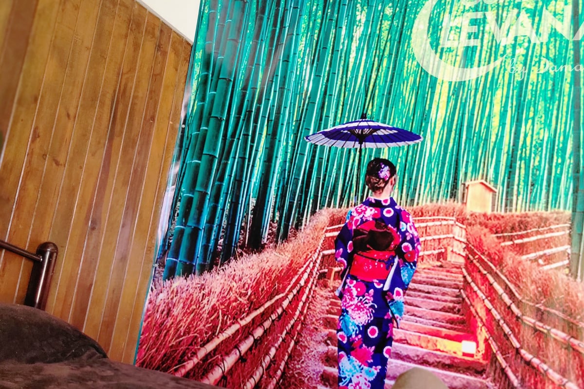 A vibrant wall mural depicts a figure in a traditional kimono, walking up a bamboo-lined path. The pathway is framed by lush greenery and soft grass, with an umbrella adding a pop of color. The wooden wall forms a cozy juxtaposition beside the mural.