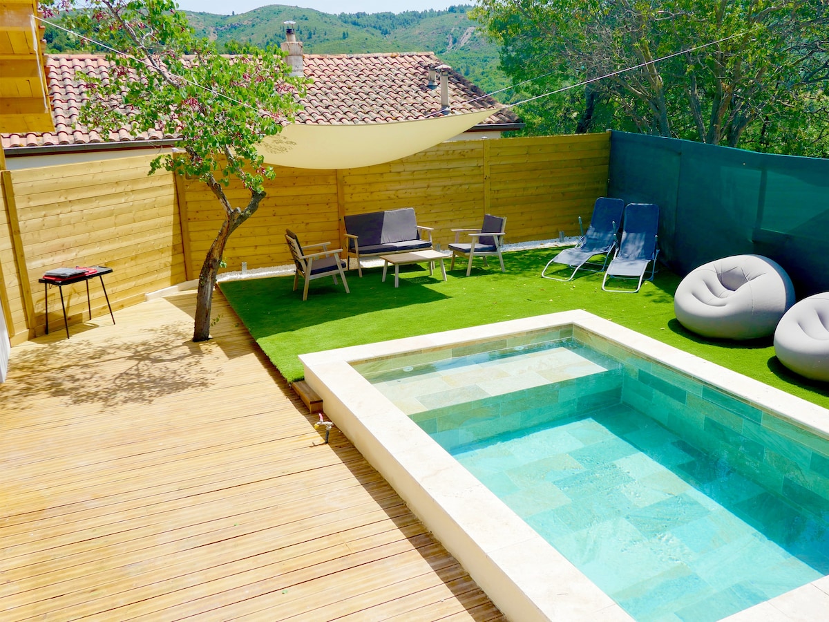 An outdoor space features a stone pool surrounded by well-maintained grass and wooden decking. Lounge chairs and bean bag seating are arranged nearby under a shaded area. A table for two sits in a corner, complemented by trees providing natural greenery.