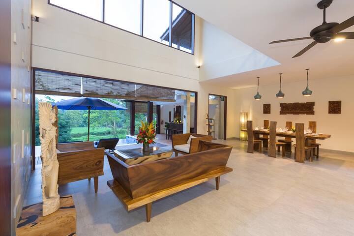 Spacious living area with high ceilings and a wall of windows overlooking lush greenery. Modern sofa and unique wood benches create a comfortable and stylish space.