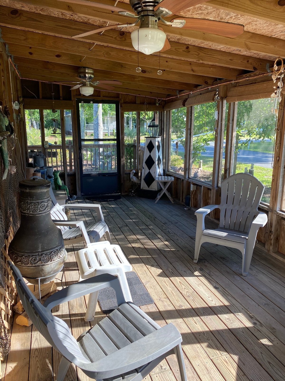 A screened front porch features wooden flooring and overhead fans, surrounded by windows allowing natural light. Several gray chairs are arranged for seating. A tall, decorative item stands against the wall, complementing the rustic charm of the space.