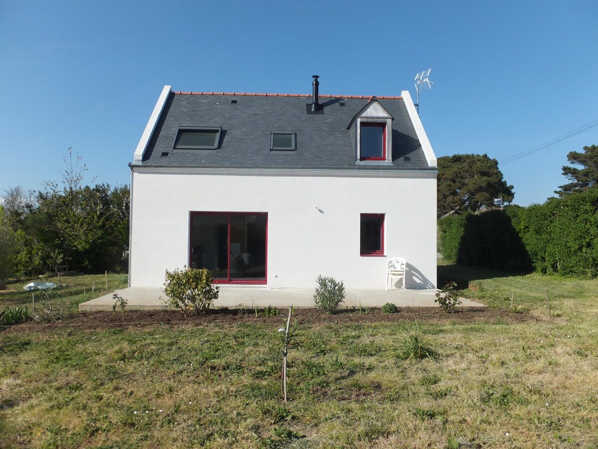 The exterior of the house is shown, featuring a modern design with white walls and a sloped roof. Large glass doors lead to the garden, which is bordered by low vegetation. A few chairs are visible on the terrace, inviting relaxation in the open space.
