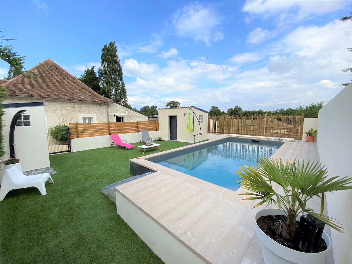A well-maintained pool area is surrounded by lush green grass, featuring a rectangular pool with clear blue water. Sun loungers are positioned on the deck, and a vibrant umbrella adds a pop of color. A rustic building is visible in the background, contributing to a serene outdoor setting.