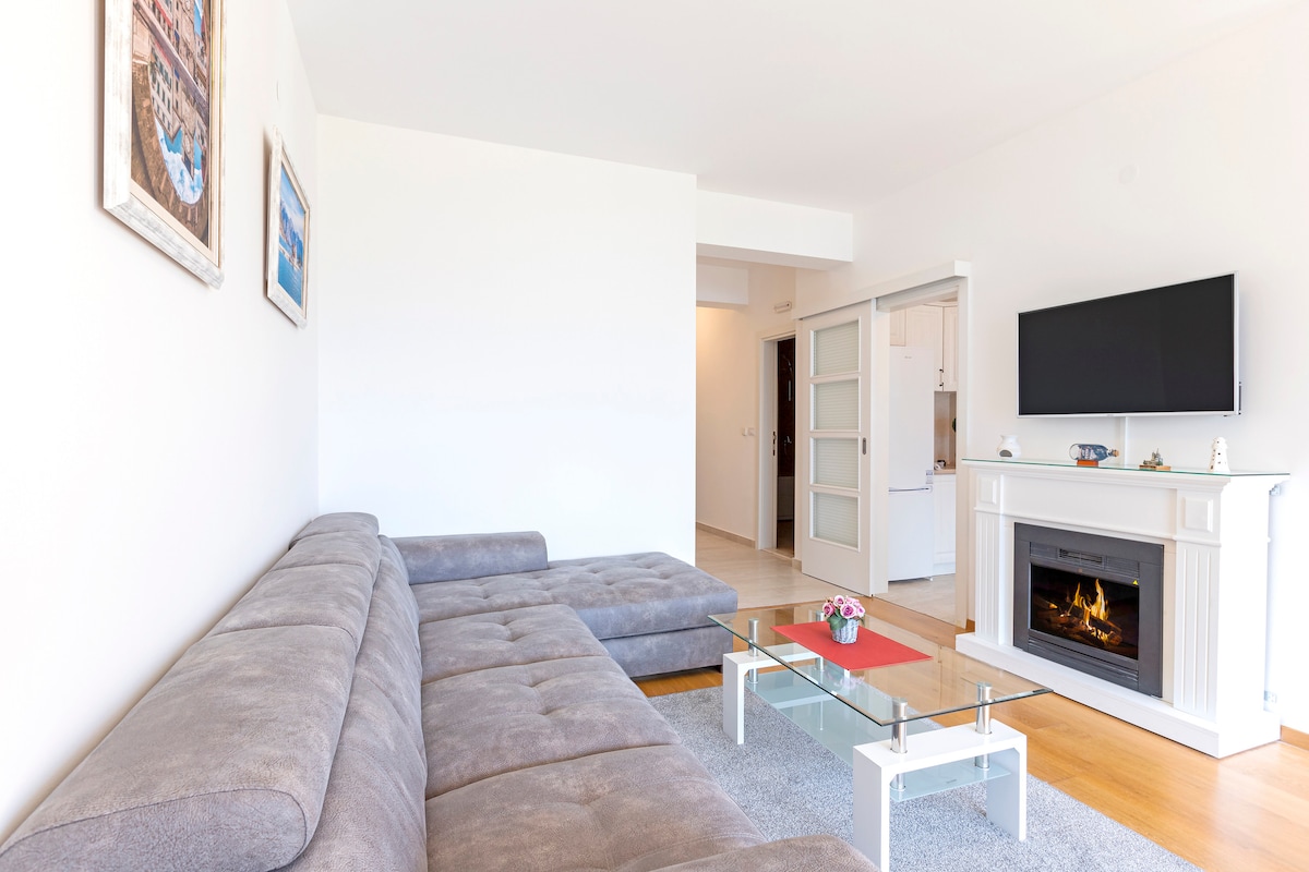 A spacious living room is highlighted by a gray sectional sofa and a glass coffee table. A modern TV is mounted above a white fireplace. Natural light fills the area through the nearby doorway, leading to the kitchen.