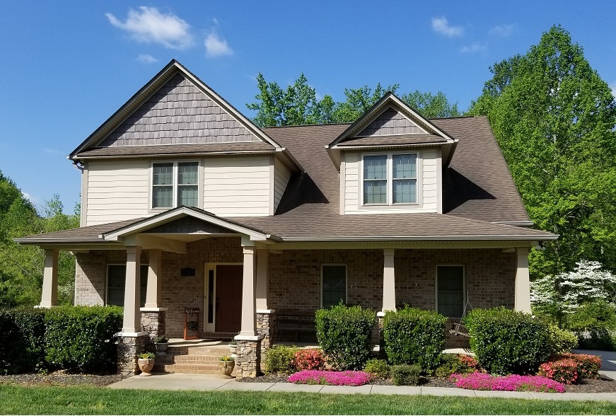 A two-story home is set amongst lush greenery, featuring a covered front porch supported by columns. Colorful flower beds line the pathway, while large windows allow natural light into the living spaces. The roof has an asymmetrical design, creating visual interest against the clear sky.