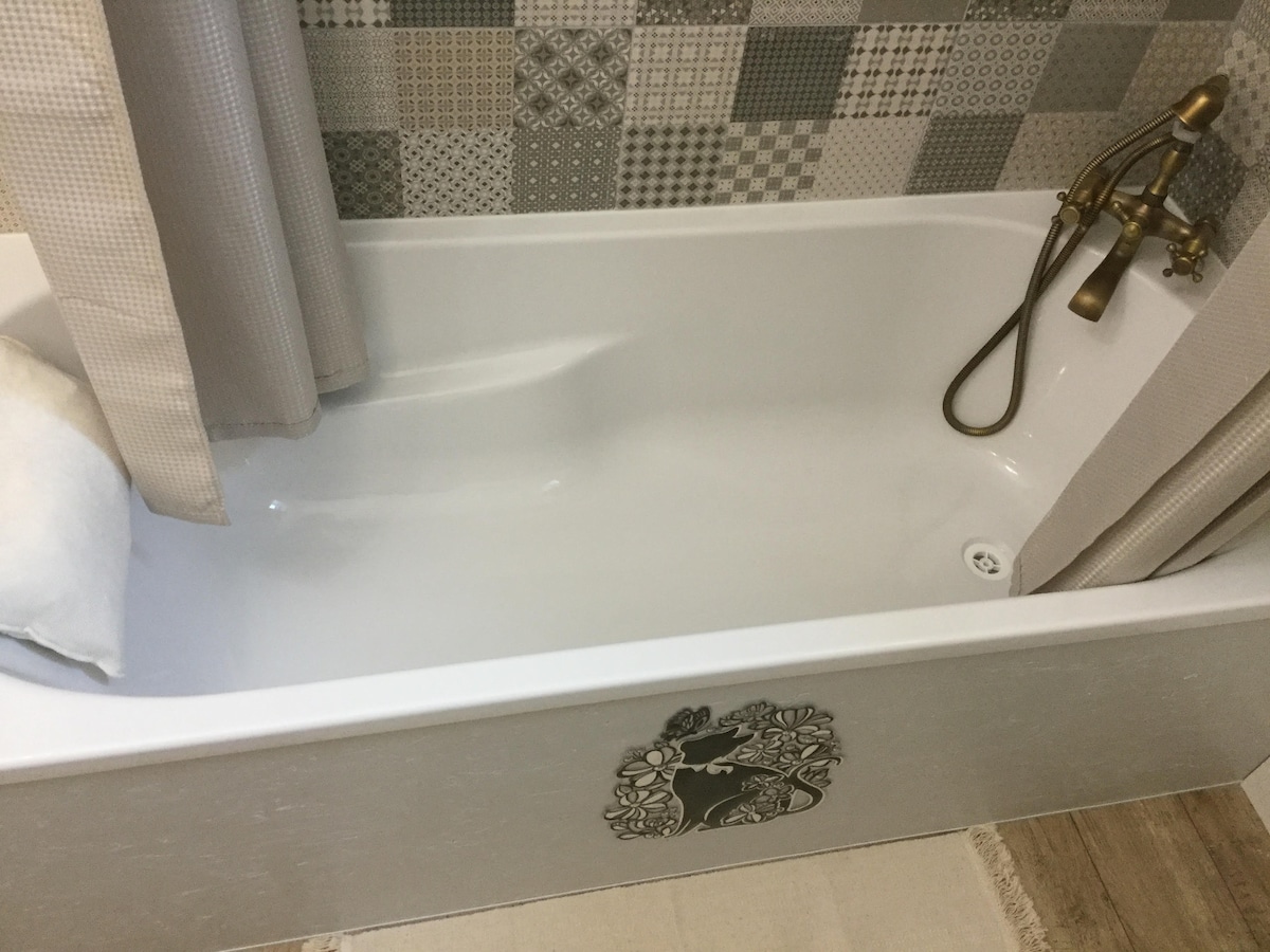 A clean bathtub is displayed against a patterned tiled wall, showcasing a blend of neutral tones. A decorative motif is featured on the side of the tub. Soft curtains frame the space, offering privacy while a light-colored towel rests on the edge.