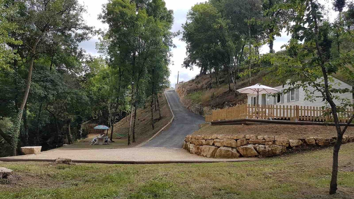 A gravel pathway leads up a gentle slope, bordered by a variety of trees. A well-maintained road ascends, with a white dwelling visible in the distance. A stone seating area is positioned on the left, while an umbrella provides shade nearby.