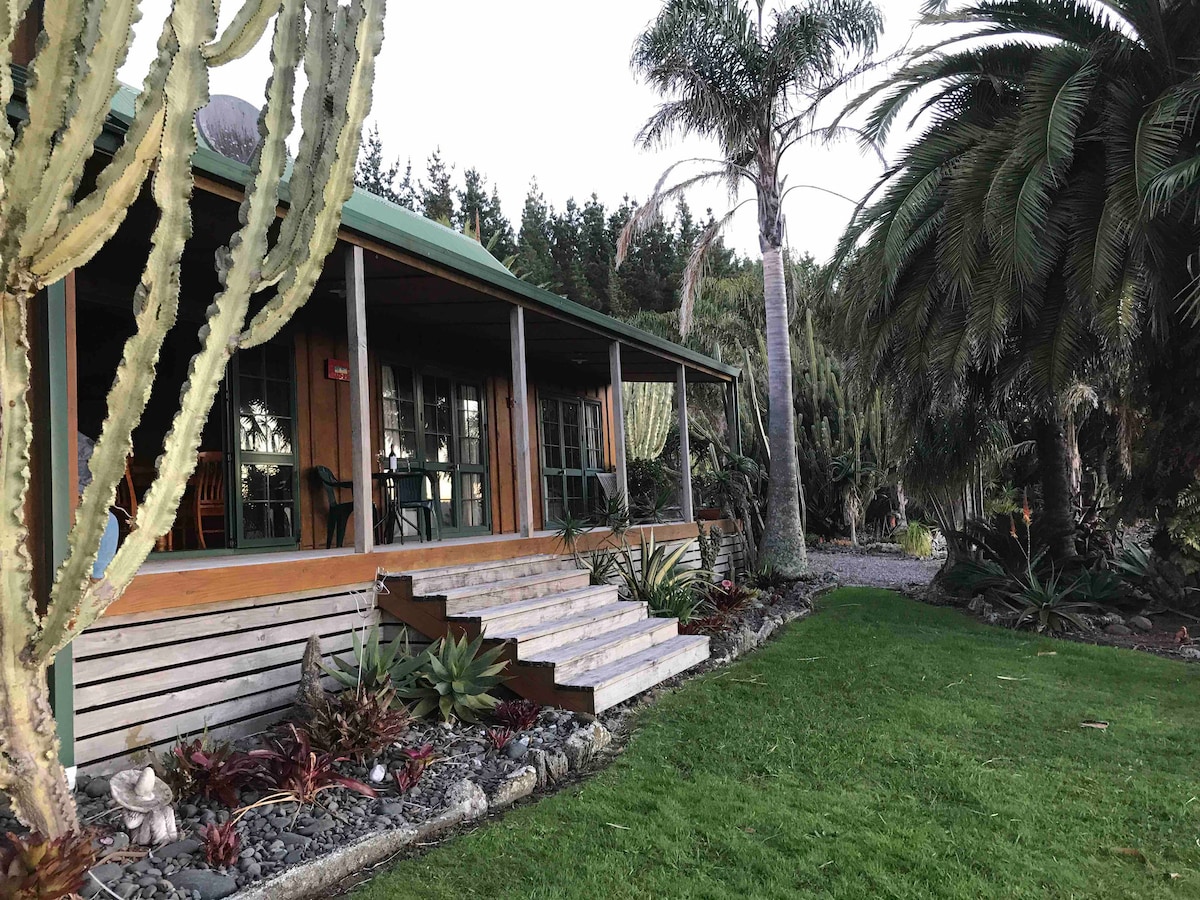 The exterior showcases a charming house surrounded by lush greenery and tropical plants, including cacti and palm trees. A set of steps leads to a covered porch with large windows, allowing views of the landscaped yard and the tranquil rural setting.