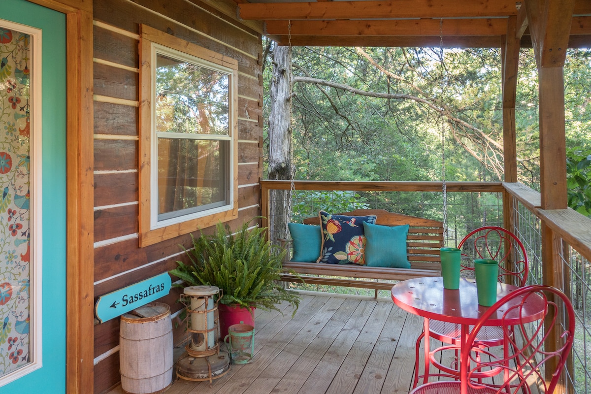 A covered porch features a swing and two red metal chairs around a small table. Bright throw pillows add color to the swing, while a potted fern enhances the natural feel. The scenery includes lush greenery seen through the railing, creating a connection to the outdoors.