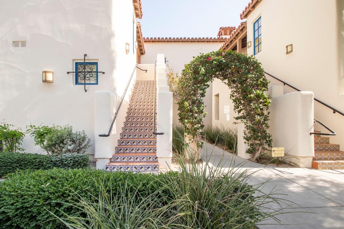 The exterior features a charming staircase adorned with colorful tiles, leading to an archway decorated with lush greenery. Neat landscaping surrounds the pathway, adding to the inviting appearance of the entrance.