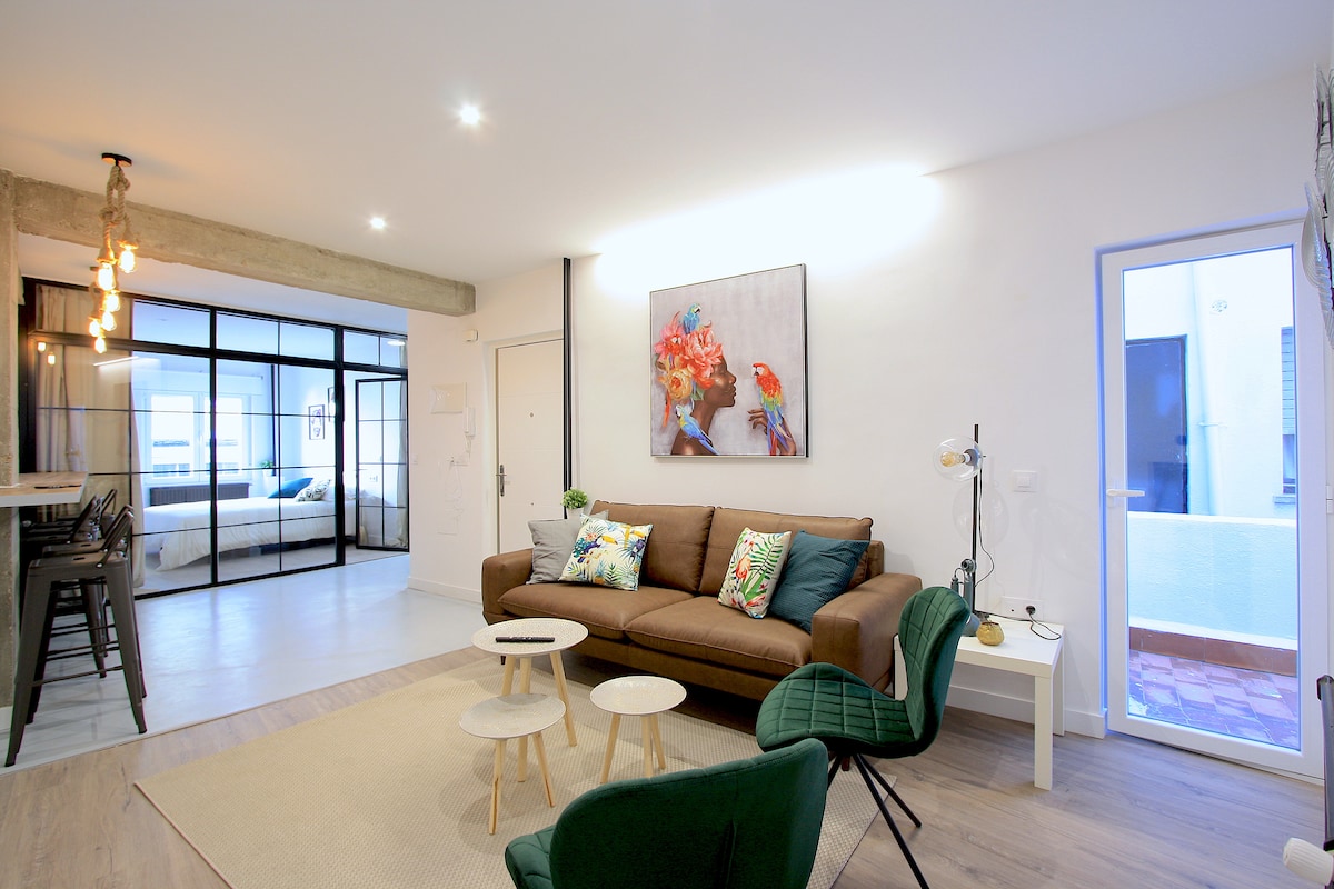 A spacious living area is highlighted by a comfortable sofa adorned with decorative cushions. Two small tables are positioned on a light-colored rug, while a large artwork featuring vibrant flowers and birds leans against the white wall. Natural light enters through a nearby glass door.