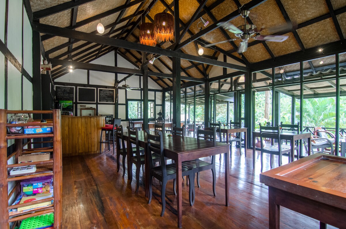 The dining area features a spacious layout with wooden tables and chairs arranged for multiple guests. Large windows allow for natural light and views of the surrounding greenery. A rustic ceiling with exposed beams and a ceiling fan adds to the inviting atmosphere.