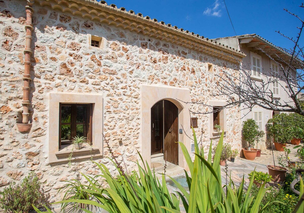 The exterior of a stone house showcases traditional architecture with a large arched entrance. Surrounding vegetation includes various potted plants and greenery, adding a touch of nature. Bright blue skies provide a contrast to the warm tones of the stone facade.