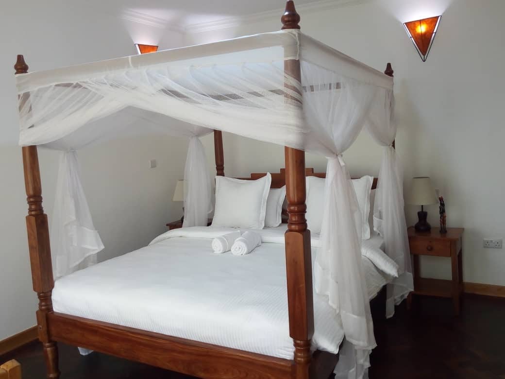 A spacious bedroom features an over-sized four-poster bed draped with sheer white fabric. Soft white bedding and neatly rolled towels are arranged elegantly on the bed. Two bedside tables with lamps flank the bed, complemented by textured wall sconces.