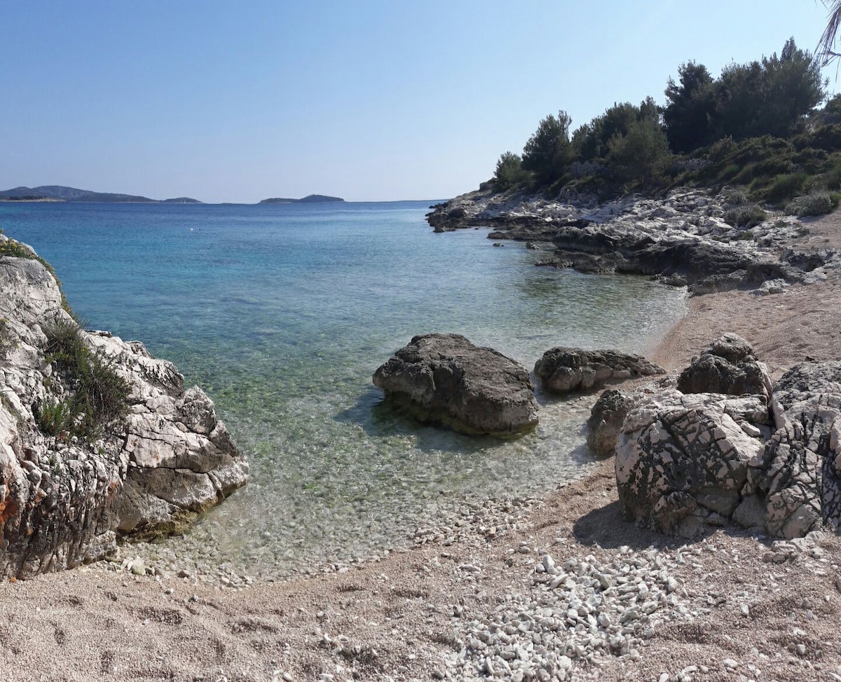 A serene beach scene features smooth, rocky formations extending into calm, clear water. The sandy shoreline is dotted with small pebbles, and gentle waves lap against the rocks. Lush greenery lines the background, contributing to a tranquil atmosphere.