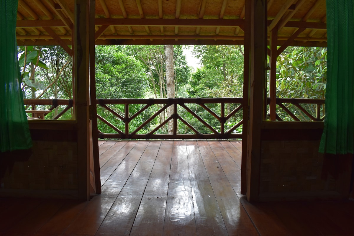 The spacious deck features polished durian wood floorboards and large openings framed by wooden railings. Lush greenery is visible through the openings, creating a seamless connection with the surrounding nature.