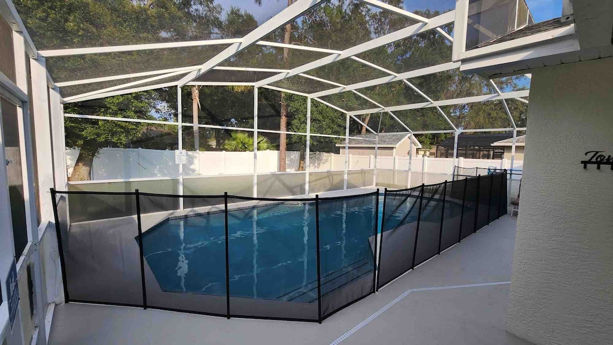A newly renovated pool area is enclosed by a protective screen, providing a safe environment. The pool is visible, surrounded by a spacious deck area adorned with light-colored flooring. Lush greenery can be seen in the background, offering a serene outdoor setting.