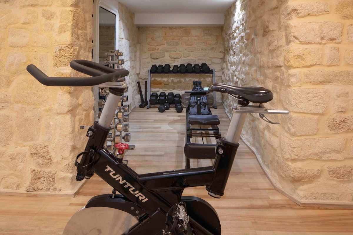A fitness area features a stationary bike prominently in the foreground, with a backdrop of stone walls. Various weights and equipment are neatly arranged, creating a dedicated workout space suited for guests looking to maintain their fitness routine.