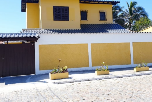 A two-story yellow house is depicted with a tiled roof and decorative windows. Surrounding the property, planters filled with green plants line a stone-paved path. A wooden gate provides access, contributing to the private and tranquil appearance of the residence.