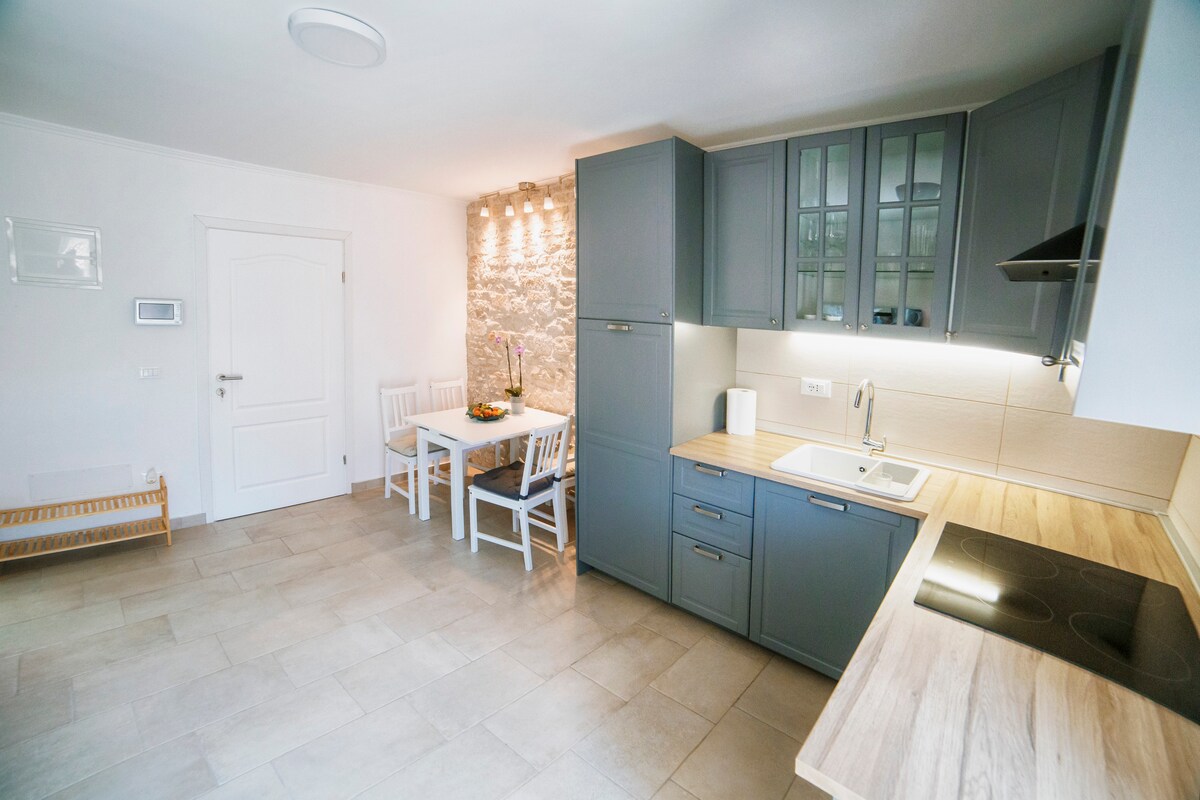 A modern kitchen and dining area are featured, showcasing gray cabinetry and a light wood countertop. A dining table with white chairs accommodates up to four people. Soft lighting highlights a stone wall, enhancing the overall bright and functional space.
