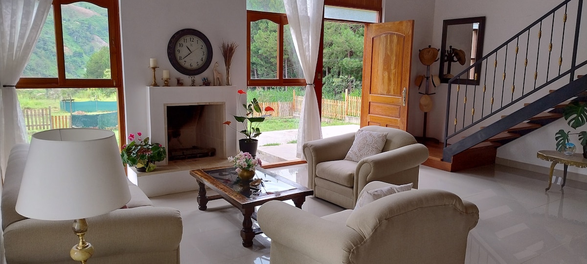 A cozy living area features soft, light-colored sofas arranged around a wooden coffee table, which holds a decorative plant. Large windows allow natural light to fill the space, showcasing views of the surrounding greenery. A fireplace adds to the warmth of the atmosphere.