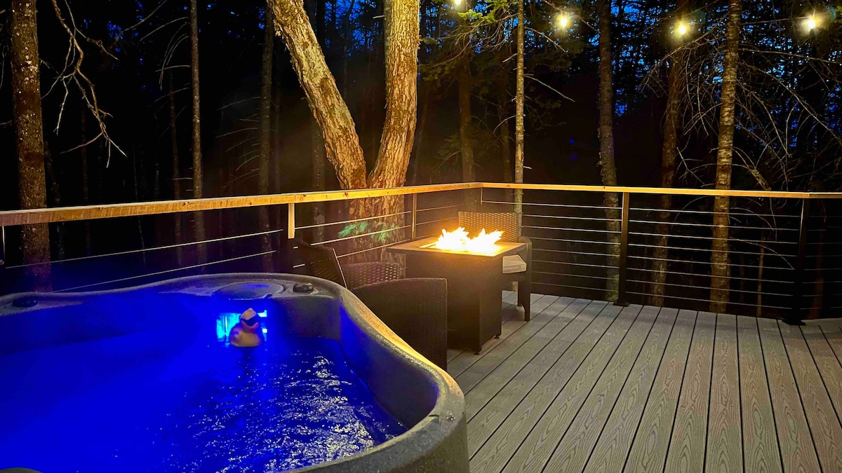 A private deck features a hot tub with soft blue lighting, nestled among trees. A propane fire table and comfortable seating provide space for relaxation. Strings of lights create a warm ambiance, enhancing the serene outdoor setting during twilight.