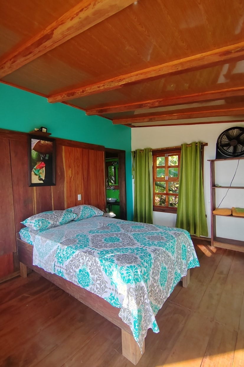 A bright sleeping area features a spacious bed covered with a patterned blanket. The walls are painted in a vibrant turquoise, complemented by wooden accents. Natural light filters through a window adorned with green curtains, and a fan is positioned nearby for comfort.