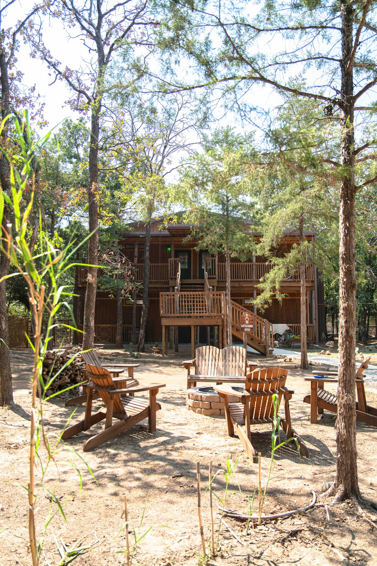 A two-story cabin is nestled among tall trees, featuring wooden decks and spacious outdoor seating. Several Adirondack chairs surround a fire pit, providing a cozy gathering spot. A path leads to the entrance of the cabin, partly concealed by lush greenery.